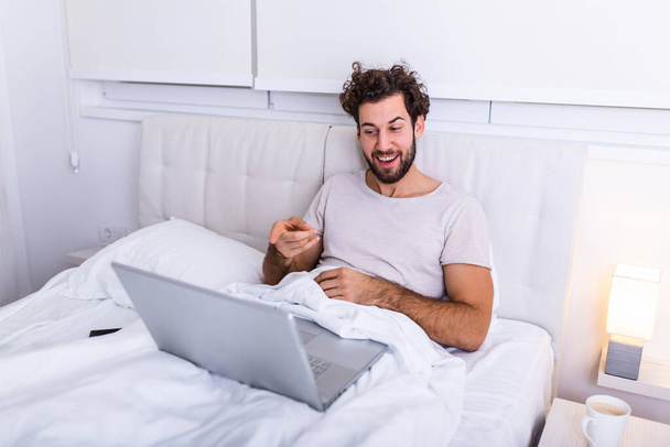 若い白人男性は朝のコーヒーを持っています,ベッドの上に座ってビデオ通話を話して座っています,本を読みます,試験の準備,サーフィンインターネットのためのラップトップコンピュータを使用します,カジュアルな家庭用服を着て. - 写真・画像