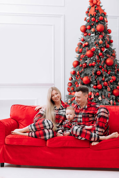 The theme of the family holiday is New year and Christmas. Young European family in the same pyjamas: mom, dad, baby boy are sitting on the red sofa by the festive Christmas tree on Christmas evening. - Photo, image