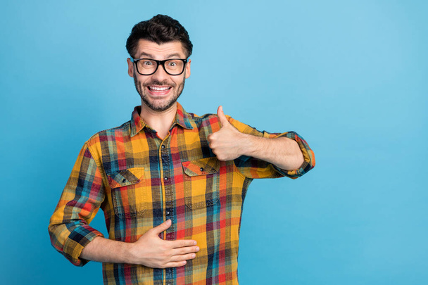Photo of satisfied cheerful positive guy in spectacles wear checkered shirt showing like arm on stomach isolated on blue color background. - Φωτογραφία, εικόνα