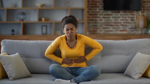 Periodenschmerz. Junge aufgebrachte Afroamerikanerin leidet unter schrecklichen Bauchschmerzen, schmerzhaftem Bauch und runzeligem Gesicht, sitzt zu Hause auf dem Sofa, Zeitlupe, freier Raum - Filmmaterial, Video