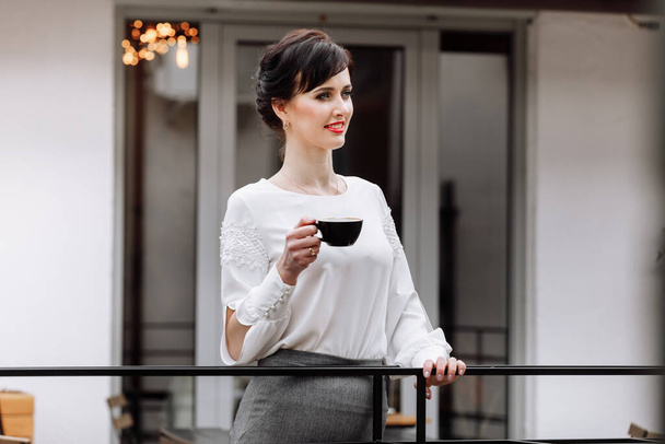 Young beautiful woman holding a cup of coffee or tea in cafe, restaurant. Brunette girl is drinking hot drinks, looking in camera outdoors on terrace and having lunch break during work. copy space. - Zdjęcie, obraz