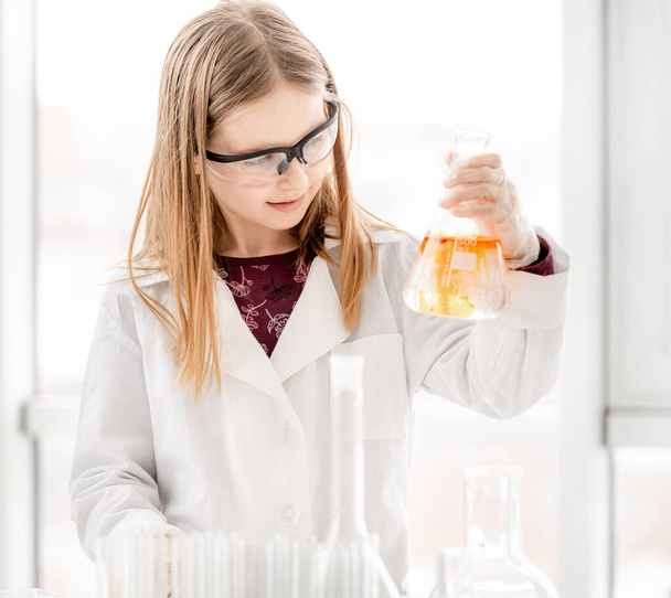 Girl doing scientific chemistry experiment wearing protection glasses and holding tube. Schoolgirl with equipment and chemical liquids on school lesson - Fotografie, Obrázek