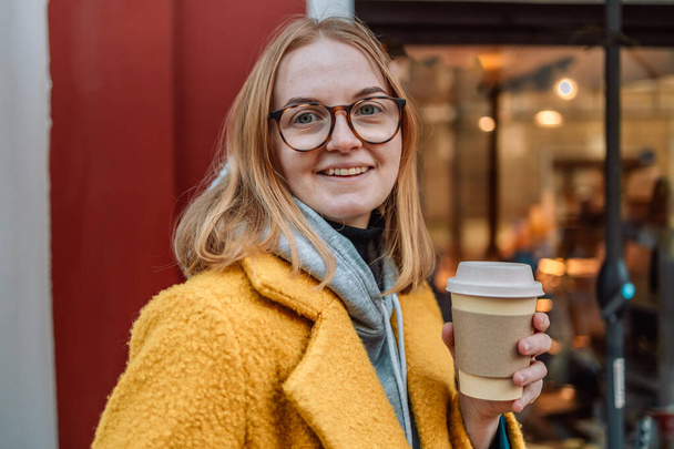 Красива і молода дівчина в пальто і шарфі сидить в міському кафе. Жінка п'є каву і сміється. Осінь
 - Фото, зображення