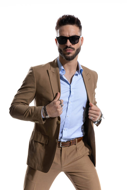 knappe serieuze man met open kraag shirt regelen bruin pak en poseren op witte achtergrond in studio - Foto, afbeelding