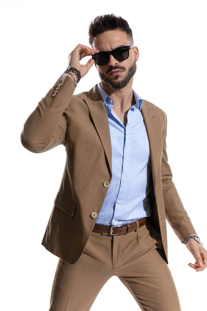 портрет сексуального бородатого бізнесмена в коричневому костюмі, що регулює сонцезахисні окуляри і позує перед білим тлом в студії
 - Фото, зображення