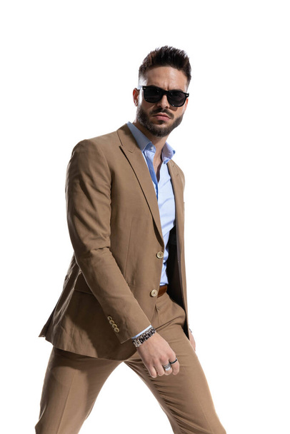 arroganter junger Mann im braunen Anzug, sein Hemd aufgelegt, mit Sonnenbrille, die cool vor weißem Hintergrund im Studio posiert - Foto, Bild