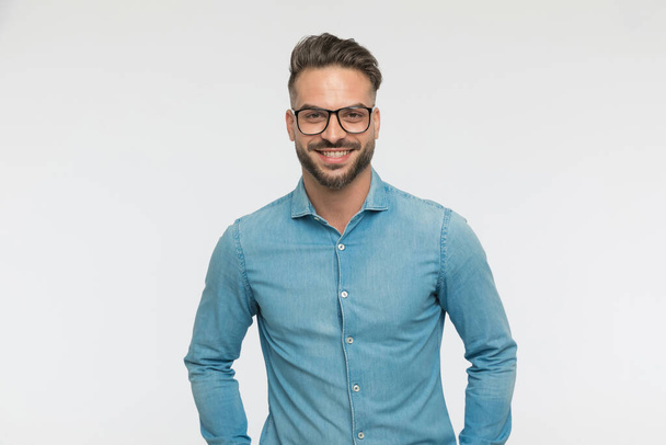 sexy casual guy dragen bril, glimlachen naar de camera en poseren tegen grijze achtergrond - Foto, afbeelding