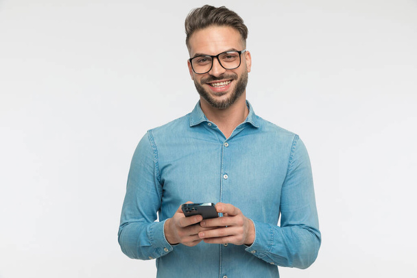 joven hombre casual feliz de charlar por teléfono con su amigo y sonriendo amplia sobre fondo gris - Foto, Imagen