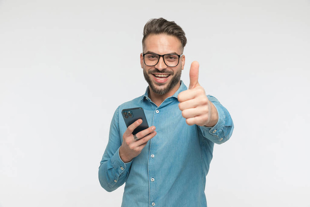 handsome casual man texting on his phone then giving a thumbs up and smiling at the camera  - Foto, Bild