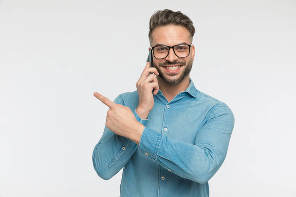 smiling casual man pointing to the side and talking on the phone against gray studio background - Fotoğraf, Görsel