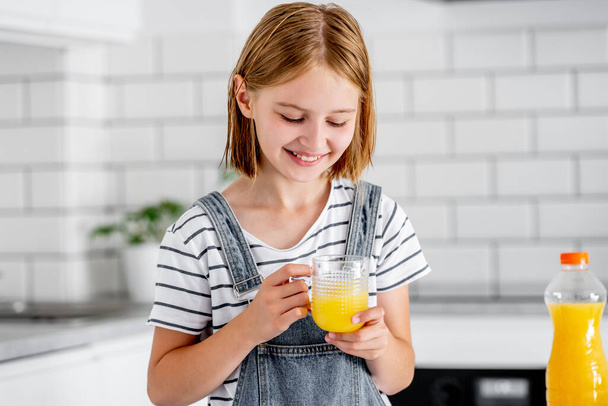キッチンで笑っているガラスの中にオレンジジュースを持つ18人の女の子。ビタミンフルーツドリンク付きの可愛い子供は家で幸せ - 写真・画像