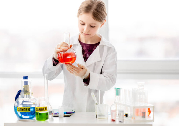 Smart girl during scientific chemistry experiment wearing protection glasses, holding bottle with orange liquid. Schoolgirl with chemical equipment on school lesson portrait - Valokuva, kuva