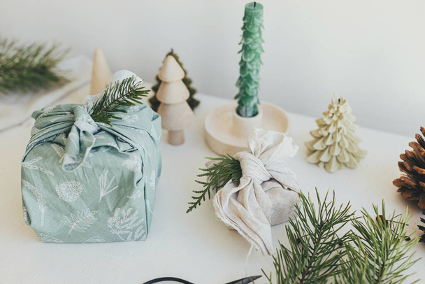 Furoshiki gift wrapping. Stylish wrapped gifts in linen fabric on white rustic table with eco friendly wooden toys, fir branches, candle. Zero waste Christmas concept. Happy Holidays! - Foto, Bild