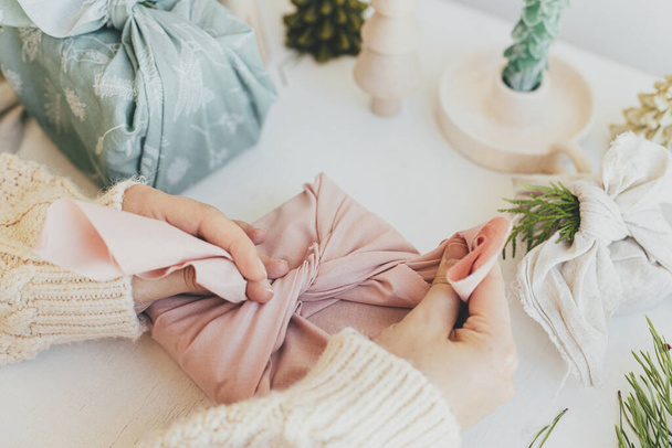 Furoshiki gift wrapping. Hands wrapping gift box in pink linen fabric on white wooden table with eco friendly wooden toy, fir branch, candle. Zero waste Christmas concept. Winter preparations - Φωτογραφία, εικόνα