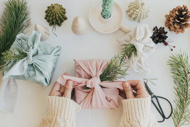 Zero waste Christmas concept. Hands holding gift box wrapped in pink fabric with fir branch on white wooden table with eco friendly decor and candle. Furoshiki gift wrapping. Eco winter holiday - Фото, изображение