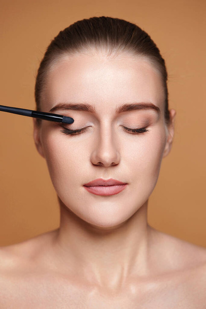 young woman applying eyeshadows on the eyelids with make-up brush on beige background. - Photo, image