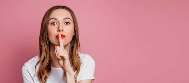 Portrait of a girl covering her mouth with her finger isolated over pink background in studio. Woman hushing. web banner, copy space - Fotografie, Obrázek