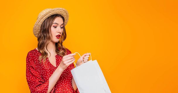 Kırmızı bir yaz elbisesi ve kırmızı dudaklı hasır şapka giyen şaşkın kız, turuncu arka planda izole edilmiş bir alışveriş çantasına bakıyor. İnternet afişi. İnternet afişi. Boşluğu kopyala - Fotoğraf, Görsel