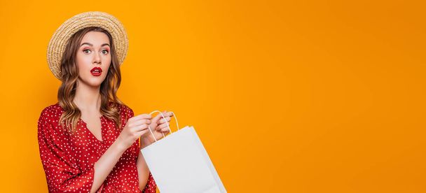 Portrait of a shocked surprised girl dressed in a red dress and straw hat with red lips holding shopping bags and looking at camera isolated over orange background. web banner. copy space - Foto, immagini