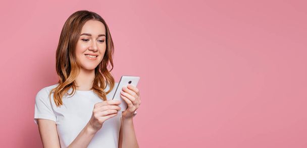 Ritratto di una giovane donna aucasica felice in una t-shirt bianca con cellulare isolato su sfondo rosa, studentessa dai capelli ricci ondulati tiene lo smartphone e legge un messaggio, copia spazio, banner web - Foto, immagini