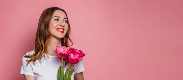 Портрет красивої молодої жінки в білій футболці, що тримає великий букет тюльпанів, ізольованих на рожевому фоні. веб-банер. копіювання простору
. - Фото, зображення