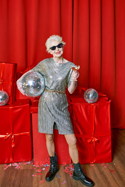 Senior élégante femme élégante avec un verre de vin mousseux sur fond rouge. Fête, mode, célébration, concept anti-âge  - Photo, image