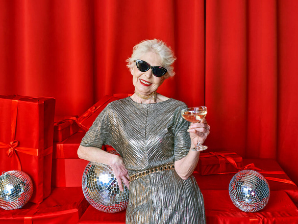 赤を基調としたスパークリングワインのグラスを持つ上級スタイリッシュなエレガントな女性。パーティー、ファッション、お祝い、アンチエイジングの概念  - 写真・画像
