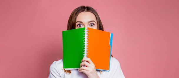 Shocked young girl student hides her face behind a notebook and surprised holds an exercise book in her hands isolated over pink background. web banner. copy space - Photo, Image