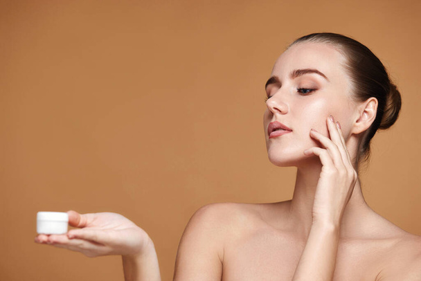 charmante vrouw aanbrengen van huidverzorging crème op haar gezicht en het houden van pot cosmetische product op beige achtergrond. kopieerruimte - Foto, afbeelding
