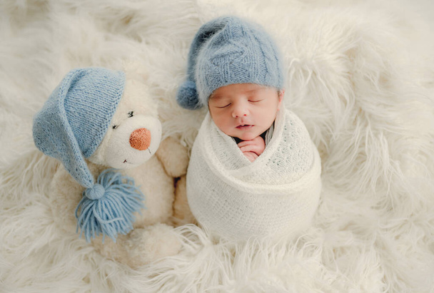 Newborn baby boy with knitted snowman toy sleeping swaddled in fabric on white fur. Infant child kid Christmas and New Year holiday portrait - Фото, изображение