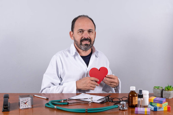 Портрет старшего зрелого врача, держащего форму сердца, смотрящего в камеру и улыбающегося - Фото, изображение