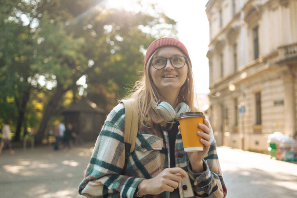 Glückliches Mädchen mit Hipster-Brille, das Musik hört, während es zur Arbeit geht. Fröhliche Frau trinkt Kaffee im Freien, glücklich lächelndes europäisches Mädchen geht - Foto, Bild