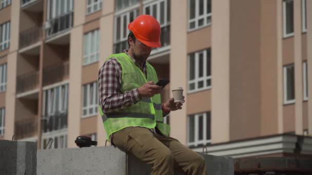 Builder si rilassa seduto su elementi di costruzione sul posto di lavoro, bere caffè e navigare in Internet su smartphone. Architetto in casco rilassarsi al lavoro e bere caffè anche andare con il telefono in mano.  - Filmati, video