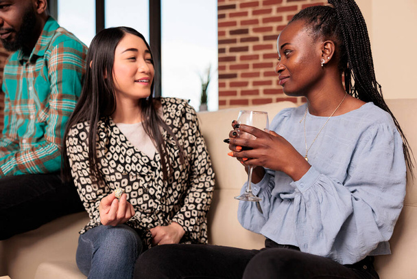 Африканська доросла жінка, яка слухає, чує розмову друга-азіата, розмовляє, п'є вино, їсть закуски в квартирі друга по вихідних.. - Фото, зображення