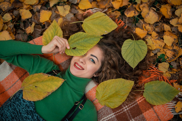 Mladá žena si užívá podzimní počasí. Kudrnatá dívka s otevřenýma očima ležící v podzimním listí, držící se za ruku a usmívající se. Podzimní nálada - Fotografie, Obrázek