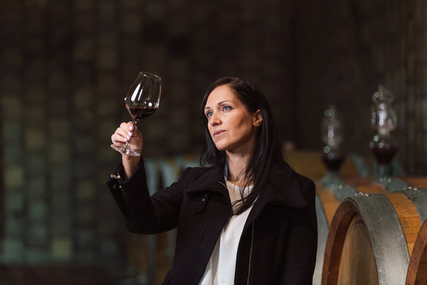 Woman in the wine cellar with barrels in background drinking and tasting wine. - Foto, afbeelding