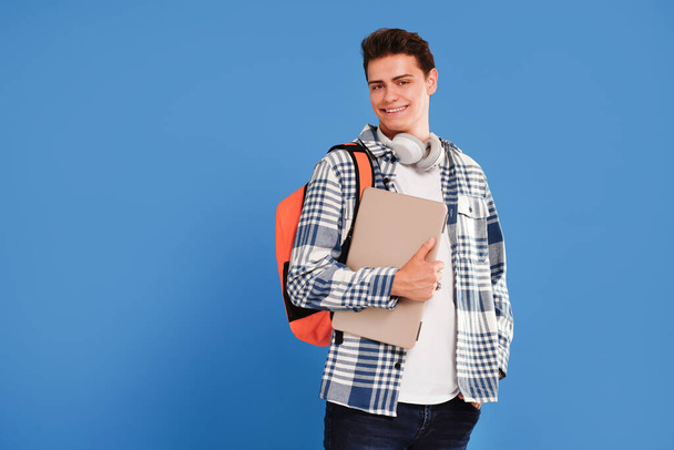 Young happy man student in casual clothes, with backpack and laptop, isolated on studio blue background. Education concept. Copy space - Фото, изображение