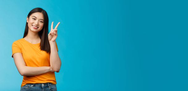 Optimistic cute tender asian young millennial girl send positive vibes, tilt head show peace, victory sign, smiling broadly, wish fantastic holidays, stand blue background joyful upbeat. - Photo, Image
