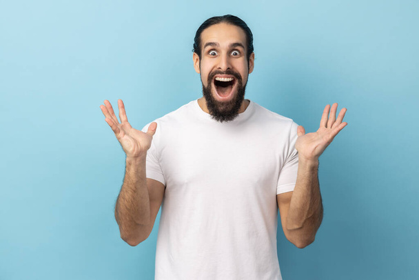 Portrait of man with beard wearing white T-shirt standing with mouth open in surprise, has shocked expression, keeping hands raised. Indoor studio shot isolated on blue background. - Foto, Imagem