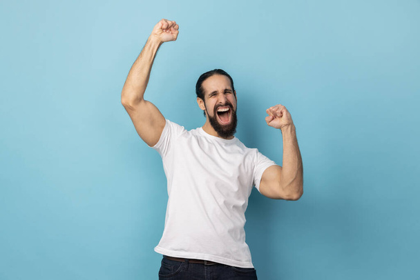 Portrait of excited man with beard wearing white T-shirt expressing winning gesture with raised fists and screaming, celebrating victory. Indoor studio shot isolated on blue background. - Zdjęcie, obraz