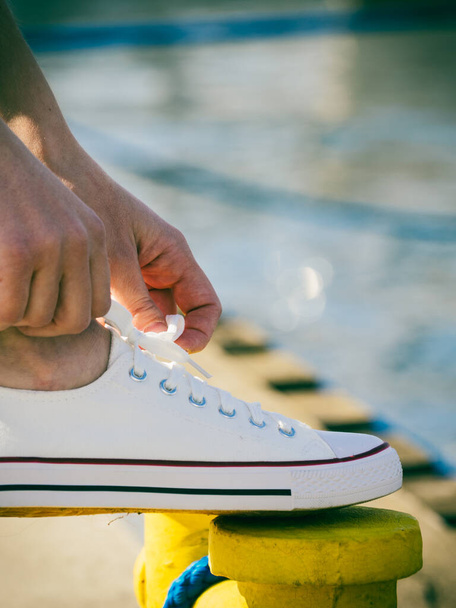 Person adjusting, tying up laces in white sneakers, casual shoes on yellow bollard pin. Outdoor footwear perfect for warm weather. - Фото, зображення