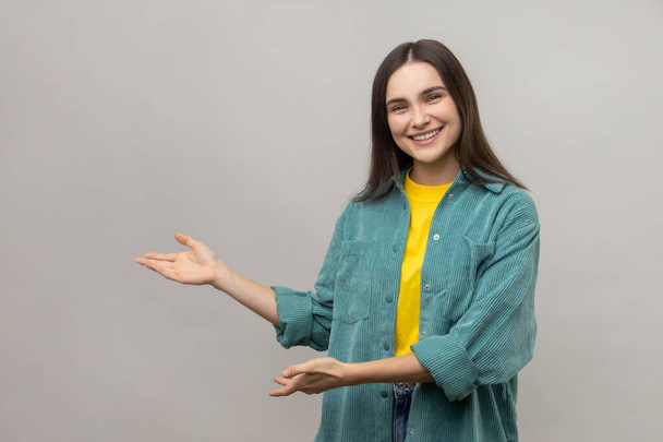 Portrait of smiling woman standing with wide raised arms and welcoming or sharing smth, looking at camera, wearing casual style jacket. Indoor studio shot isolated on gray background. - Photo, image