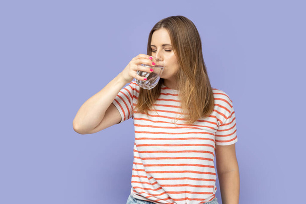 Portrait of thirsty attractive beautiful young adult blond woman wearing striped T-shirt drinking water from glass, healthy eating. Indoor studio shot isolated on purple background. - Photo, Image