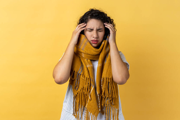 Portrait of sick ill woman with dark wavy hair wrapped in warm scarf, suffering headache, massaging her temples, having flu symptoms. Indoor studio shot isolated on yellow background. - Fotó, kép