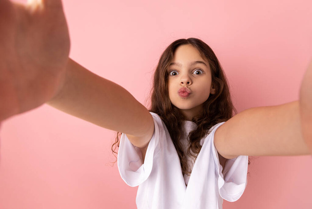 Portrait of adorable little girl wearing white T-shirt sending air kisses while taking selfie POV, flirting, expressing romantic feelings. Indoor studio shot isolated on pink background. - Фото, изображение
