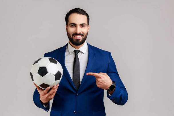 Handsome bearded businessman pointing finger at soccer ball on his hand with smiling positive expression, wearing official style suit. Indoor studio shot isolated on gray background. - Valokuva, kuva