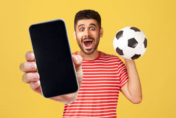 Portrait of excited surprised amazed man in red T-shirt holding and showing empty display smartphone and football ball, betting on soccer. Indoor studio shot isolated on yellow background. - Foto, immagini