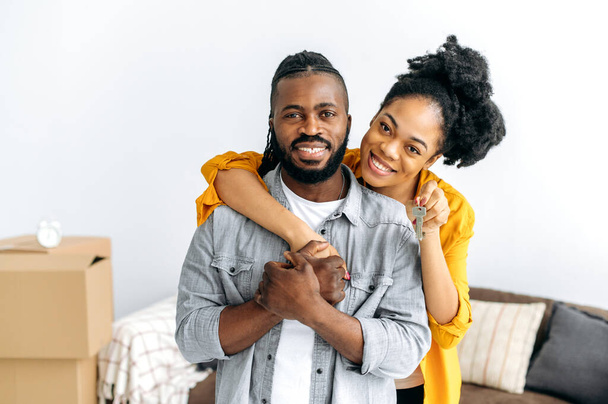 Концепція володіння. Щаслива афро-американська сім'я, чорна пара, що обіймається стоячи у вітальні, жінка, що показує ключі від своєї нової власності, вони дивляться на камеру, усміхаються - Фото, зображення
