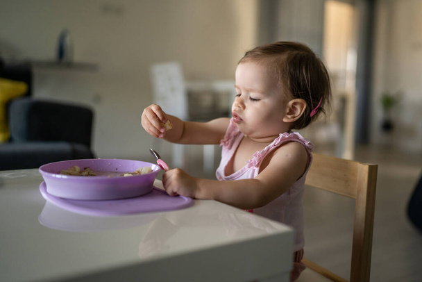 One child small caucasian toddler female baby eating at the table alone at home in room real people copy space early child development concept learning front view hold food healthy eating - Photo, Image