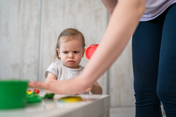 Одна маленькая кавказская девочка сердитая маленькая девочка девочка дочь сидит за столом дома несчастный или сердитый в то время как ее мать собирает или забирает игрушки взрослея детская концепция - Фото, изображение